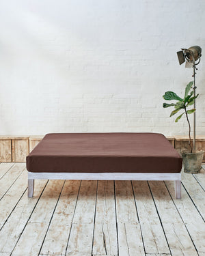 lifestyle shot of acorn brown fitted sheet, situated within a modern bedroom with rustic wooden flooring and white exposed brick walls. 